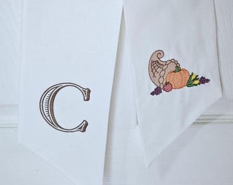 May include: White fabric door hanger with a brown embroidered letter "C" and a fall harvest cornucopia design. The cornucopia includes a pumpkin, grapes, and other harvest elements. The door hanger is hanging on a white door.
