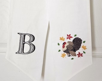 May include: White fabric door hanger with a black embroidered letter "B" on one side. The other side features an embroidered turkey surrounded by fall leaves. The door hanger is a festive decoration.