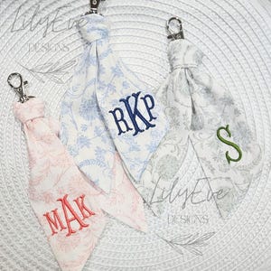 May include: Three fabric keychains with silver-tone lobster clasps. The left keychain is pink with floral patterns and the monogram MAK. The center is blue with floral patterns and RKP. The right is gray with floral patterns and the letter S.