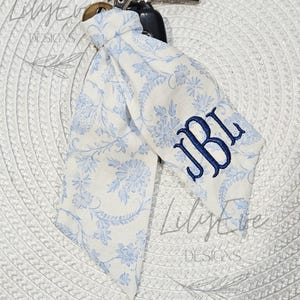 May include: A fabric keychain accessory featuring a light blue and white floral pattern with the embroidered monogram JBL in dark blue thread. The fabric is knotted onto a metal clasp attached to a string of light-colored wooden beads and a key.