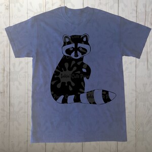 May include: A blue t-shirt with a black and white raccoon graphic. The raccoon is sitting up with its paws in the air and has a black and white striped tail.