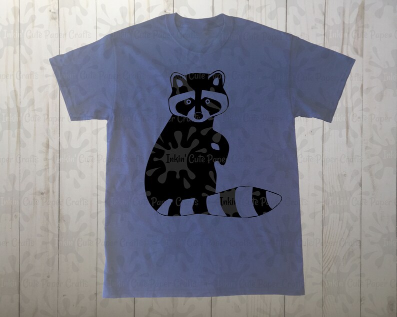 May include: A blue t-shirt with a black and white raccoon graphic. The raccoon is sitting up with its paws in the air and has a black and white striped tail.