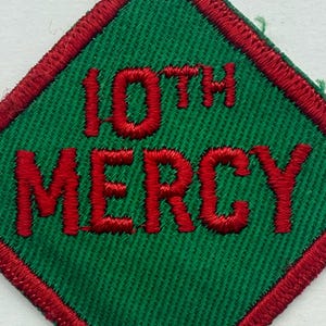 May include: A diamond-shaped patch with green fabric and red embroidered text reading "10TH MERCY". The patch has a red border, and the text is in a bold font.