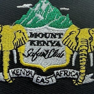 May include: A circular black embroidered patch featuring two yellow elephants flanking a central shield. The shield displays a green mountain and the text MOUNT KENYA Safari Club. A white banner below reads KENYA EAST AFRICA in black text.