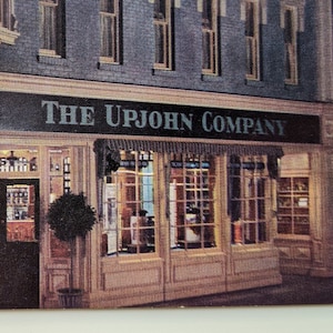 May include: A vintage postcard showing a shop front at night with the text "The Upjohn Company" above the windows. The building is brick with white trim and has a large, ornate sign with the company's name.