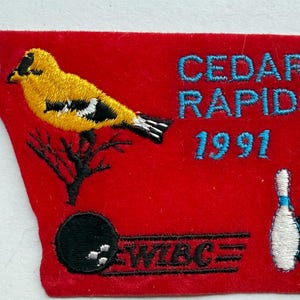 May include: A red embroidered patch shaped like the state of Iowa featuring a yellow goldfinch on a black branch, the text CEDAR RAPIDS 1991, a blue star, a black bowling ball, a white bowling pin, and the black text WIBC with horizontal lines.