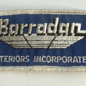 May include: A rectangular blue patch with white embroidered text and a stylized boat design. The text reads "Barradan" above "INTERIORS INCORPORATED". The patch has a white border and is likely a logo or emblem.