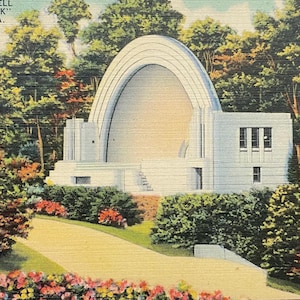 May include: A vintage postcard depicting a white band shell in a park setting. The band shell has a large arched opening and is surrounded by lush green trees and colorful flowers. The text on the postcard reads "The Band Shell in "City Park" Reading, PA."