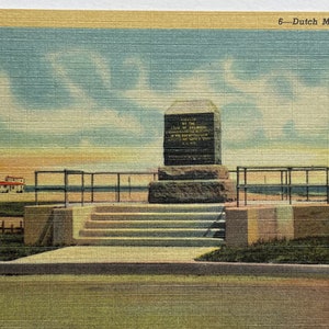 May include: A postcard depicting a stone monument with a plaque commemorating the Dutch settlement of Lewes, Delaware. The monument is situated on a grassy area with steps leading up to it. The background features a blue sky with white clouds and a view of the ocean.