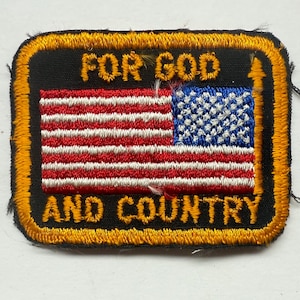 May include: A rectangular patch with a black background and a gold border. The patch features the American flag with red and white stripes and a blue field of stars. The words "FOR GOD" are above the flag, and "AND COUNTRY" are below.