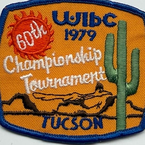 May include: A rectangular embroidered patch with a blue border on an orange background. It features a red sun, a green saguaro cactus, a brown desert landscape, and text reading WIBC 1979 60th Championship Tournament TUCSON. A ruler shows it is 10.16 cm wide.