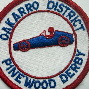 May include: Circular embroidered patch with a red and white border. The patch has the text "OAKARRO DISTRICT PINE WOOD DERBY" in blue. Inside the circle is a blue race car with a red driver.