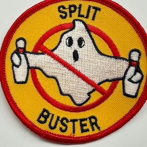May include: A circular embroidered patch with a yellow background and a red border. It features a white ghost character holding two white bowling pins. A red diagonal line crosses the ghost. The black text SPLIT BUSTER is printed on the patch.