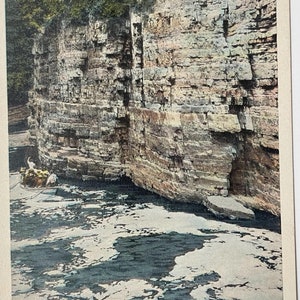 May include: A vintage postcard showing a rocky cliff face with a river flowing in front of it. The river is whitewater rapids. The text on the postcard reads "Nearing the Rapids, Ausable Chasm, N.Y."