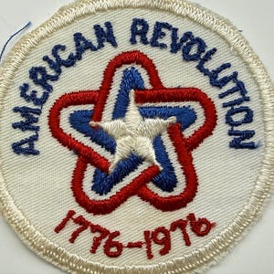 May include: A circular embroidered patch with a white background and a stitched border. It features the text AMERICAN REVOLUTION in blue, a central star design in red, white, and blue, and the red embroidered dates 1776-1976 at the bottom.