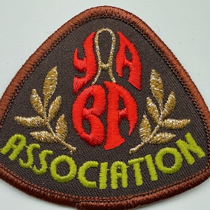 May include: A triangular embroidered patch with a dark brown background and a brown stitched border. It features red embroidered letters YABA, two gold metallic laurel branches, and the word ASSOCIATION in lime green thread at the bottom.