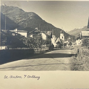 May include: A black and white vintage postcard showing a street scene in St. Anton am Arlberg, Austria. The postcard features a view of a street lined with buildings, trees, and a church steeple in the distance. The text "St. Anton y Arlberg" and "Foto Rio" are printed on the postcard.
