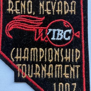 May include: A black, state-shaped embroidered patch featuring red borders and gold lettering that reads RENO, NEVADA, CHAMPIONSHIP TOURNAMENT, and 1997. The patch includes a red graphic element and the white text WIBC inside a red circular outline.