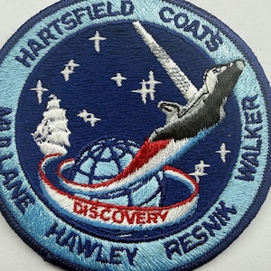 May include: A circular embroidered patch featuring a dark blue space scene with white stars, a stylized space shuttle, and a globe. The text reads HARTSFIELD, COATS, MULLANE, HAWLEY, RESNIK, WALKER, and DISCOVERY. The border is light blue.