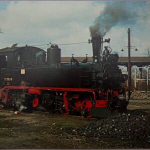 May include: Vintage black steam locomotive with red accents, billowing smoke, and the number 99 663-0. The train is parked on a gravel surface, with a pile of coal in the foreground and industrial structures in the background.