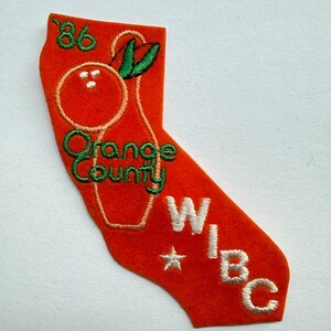 May include: An orange felt patch shaped like the state of California featuring embroidered details including the text '86', 'Orange County', and 'WIBC' in white and green thread, along with a graphic of an orange fruit and a small white star.