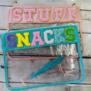 Può includere: Due pochette trasparenti con cerniera, una rosa e una turchese, con le parole "STUFF" e "SNACKS" in lettere colorate e strutturate. Le pochette hanno un fronte trasparente. La pochette turchese include un cinturino da polso turchese.