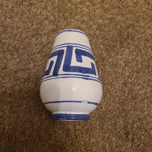 May include: White ceramic vase with blue geometric patterns. The vase has a rounded body and a slightly flared top. The blue design is a repeating pattern of angular shapes. The base and top rim are also blue.
