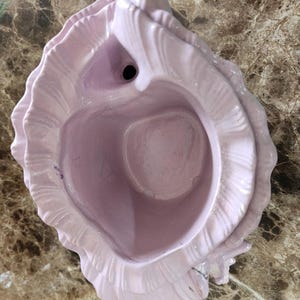 May include: A light pink ceramic seashell-shaped decorative bowl. The bowl has a central cavity and a hole. The exterior has a textured, ruffled design. The bowl is on a brown and tan marble-patterned surface.