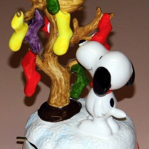 May include: A white ceramic musical figurine of Snoopy standing under a tree with a yellow bird in a nest. The tree is decorated with red, green, and yellow stockings. The base of the figurine is white with a red rim and depicts snow.