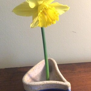 May include: A bright yellow daffodil with a green stem is displayed in a ceramic vase. The vase is triangular, with a speckled beige interior and a blue and beige exterior. The flower is in full bloom.