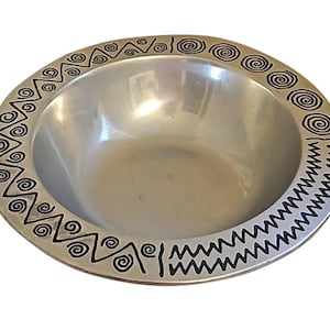 May include: A silver-colored bowl with a decorative black border featuring spiral and zigzag patterns. The bowl has a wide, shallow design, suitable for serving or display. The rim is adorned with repeating geometric shapes.
