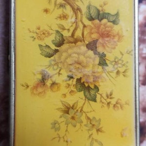 May include: A small, yellow metal box with a floral design. The box has a hinged lid and a floral pattern on the front. The flowers are yellow, white, and green.