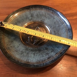 May include: A decorative plate with a teal and gold finish. The plate features a central medallion with a detailed relief design. The plate measures approximately 10 inches in diameter, as indicated by a measuring tape.