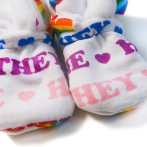 May include: Two white baby booties with rainbow trim and the words "THEY" and "HEY" printed in pink and purple. Blue heart and rainbow accents adorn the top. The booties are made of a soft, plush material.