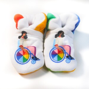 May include: A pair of white baby booties with a colorful rainbow wheel design. The booties feature a person in a wheelchair with a rainbow wheel.