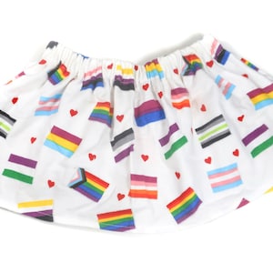 May include: A white skirt with a variety of pride flags printed on it. The flags include the rainbow flag, the transgender flag, the bisexual flag, the lesbian flag, the pansexual flag, the asexual flag, the genderqueer flag, the intersex flag, and the non-binary flag.