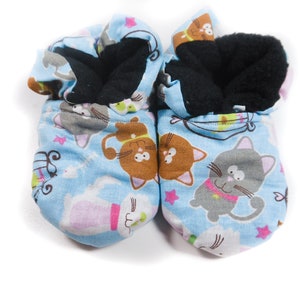 May include: A pair of blue baby booties with a cat print. The booties are lined with black fleece and have a white star pattern.