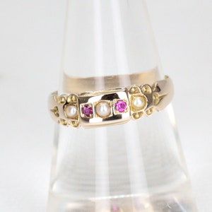 May include: A gold-toned ring featuring a rectangular central design with a pearl flanked by two vibrant pink gemstones. The ring's band is adorned with additional pearls and decorative elements, creating an elegant and vintage aesthetic.