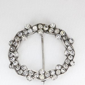 Op de afbeelding: Een zilverkleurige, ovaalvormige broche, bezet met talrijke heldere, sprankelende strass-steentjes. De broche heeft een speldsluiting aan de achterkant en staat tegen een witte achtergrond. Het ontwerp is symmetrisch en sierlijk.