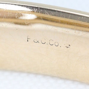 Puede incluir: Primer plano de una banda de metal dorada con la inscripci&oacute;n "F & C. Co." en el interior.