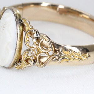 Puede incluir: Un brazalete de oro con un camafeo blanco que representa el perfil de una mujer. El brazalete tiene un dise&ntilde;o delicado y ornamentado.