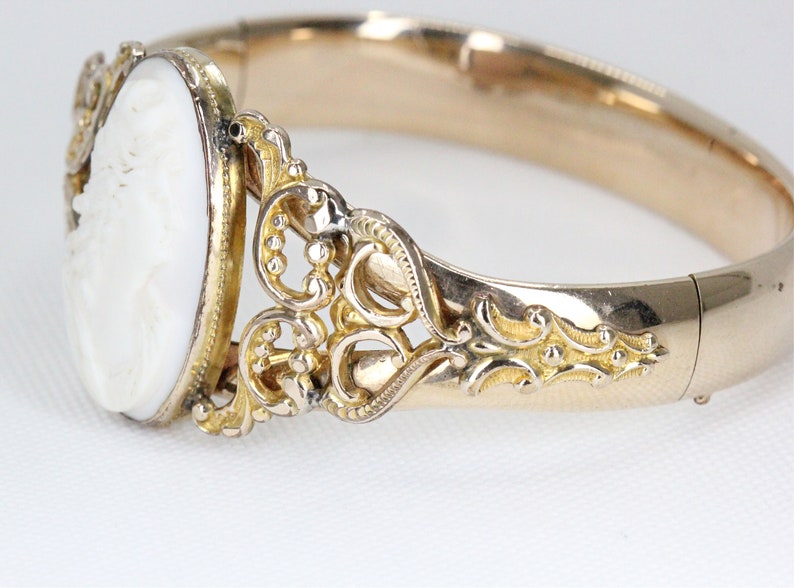 Puede incluir: Un brazalete de oro con un camafeo blanco que representa el perfil de una mujer. El brazalete tiene un dise&ntilde;o delicado y ornamentado.