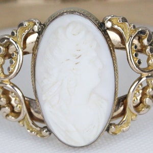 Puede incluir: Un brazalete de oro con un camafeo blanco que representa el perfil de una mujer. El camafeo est&aacute; engastado en un marco delicado y ornamentado.
