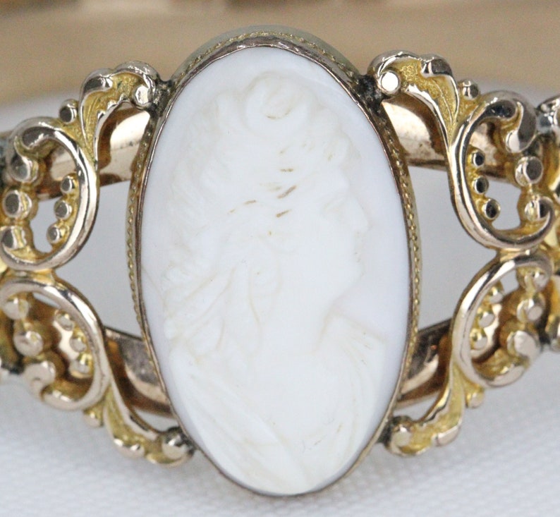 Puede incluir: Un brazalete de oro con un camafeo blanco que representa el perfil de una mujer. El camafeo est&aacute; engastado en un marco delicado y ornamentado.
