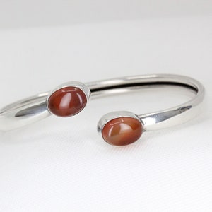 Può includere: Un bracciale a polsino argentato con due pietre preziose ovali marrone-rossastre. Il bracciale ha una finitura liscia e lucida e un design aperto. Le pietre preziose sono incastonate in una montatura a castone, aggiungendo un tocco di eleganza.