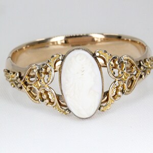 Puede incluir: Un brazalete de oro con un camafeo blanco que presenta un perfil tallado de una mujer. El brazalete tiene un dise&ntilde;o delicado y ornamentado.