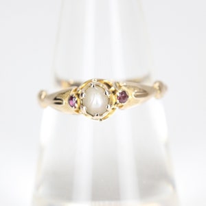 May include: A gold ring featuring a central pearl surrounded by a decorative setting. Two small red gemstones flank the pearl. The ring has an ornate, vintage design.