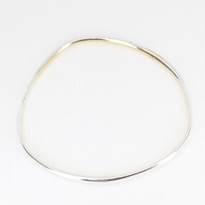 May include: A silver bangle bracelet with an irregular, organic shape. The bracelet has a smooth, polished finish and a thin profile. The inner edge has a gold-coloured accent. The bracelet is displayed against a white background.