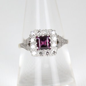 Może przedstawiać: Srebrny pierścionek z kwadratowym, fioletowym kamieniem szlachetnym otoczonym aureolą mniejszych białych kamieni.