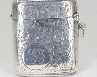 Könnte beinhalten: Eine rechteckige silberne Box mit Scharnierdeckel, verziert mit aufwendigen floralen und scrollartigen Gravuren. Ein kreisförmiges Monogramm ist mittig auf der Vorderseite angebracht. Die Box hat einen kleinen Ring an der Seite.